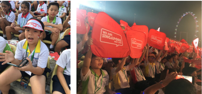 Children in white uniforms at a We Are Singapore event, seated in stands and raising red bags in the air.